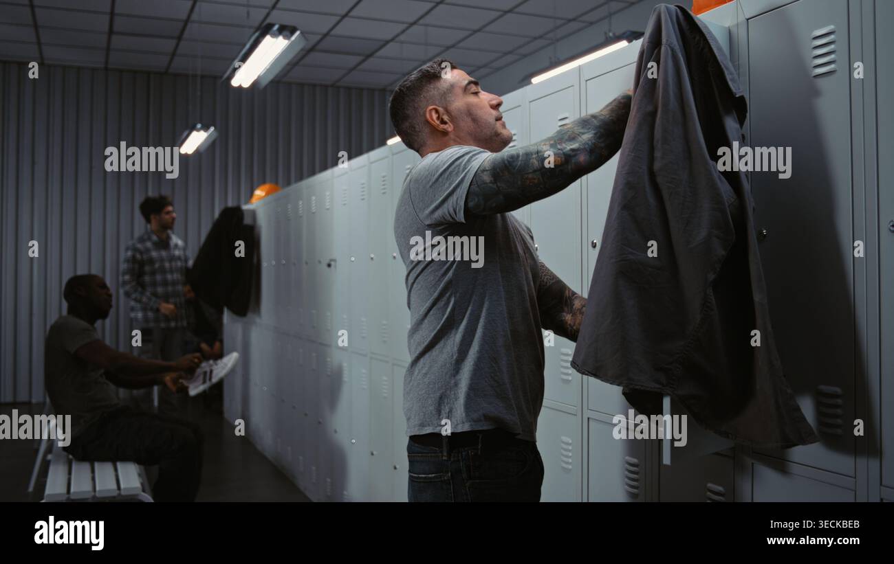 Drei verschiedene Industriearbeiter in High Visibility Gear Friendly interagieren, lachen und teilen Witze, während sie sich für ihre Schicht in der modernen Fabrik kleiden. Unternehmenskultur und Teamgeist. Stockfoto
