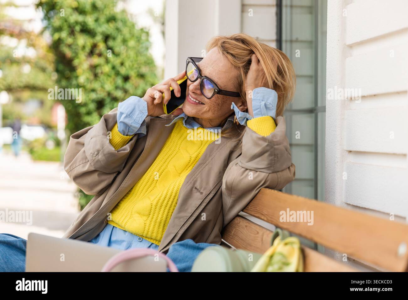 Eine stilvolle Frau mit Brille spricht auf ihrem Smartphone, während sie draußen auf einer Bank sitzt und einen entspannten Lebensstil und eine lebendige Mode in der Sonne präsentiert Stockfoto