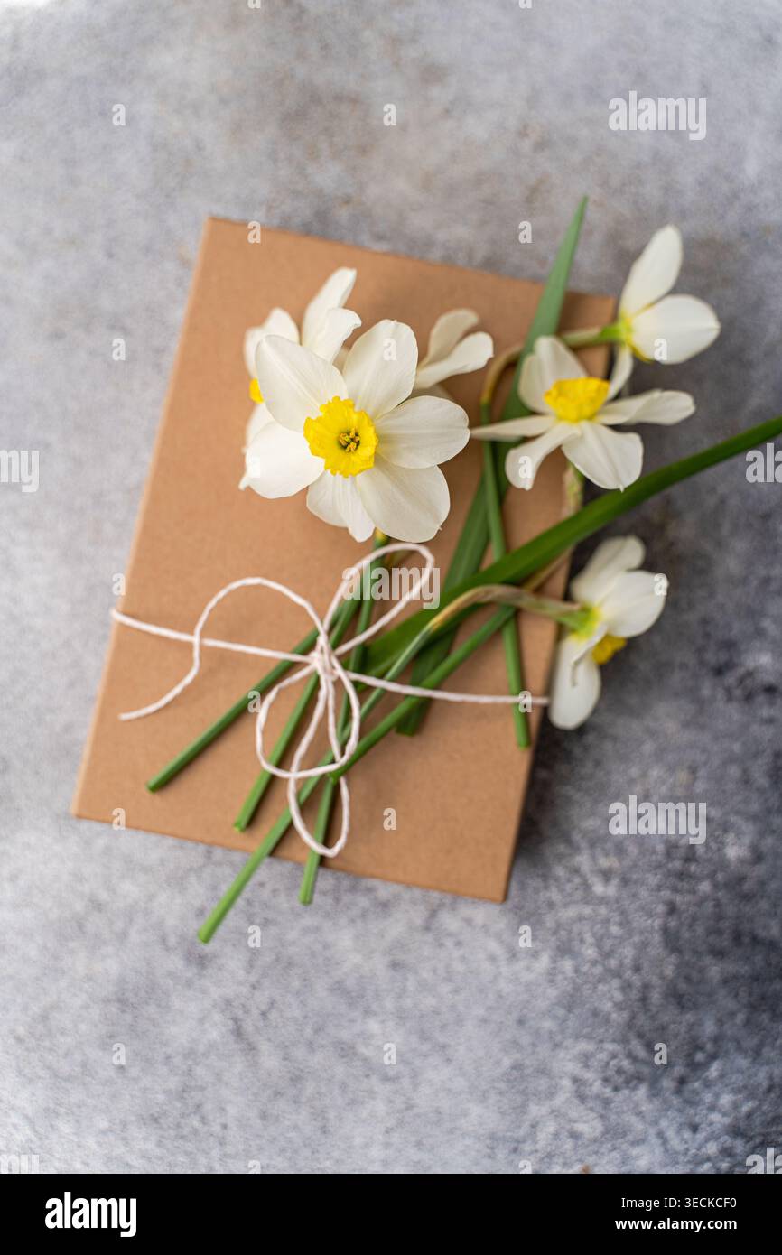 Ein Haufen zarter weißer Narzissen mit gelben Mittelpunkten liegt elegant auf einer rustikalen braunen Geschenkbox, die die Erneuerung des Frühlings und die Schönheit von nat symbolisiert Stockfoto