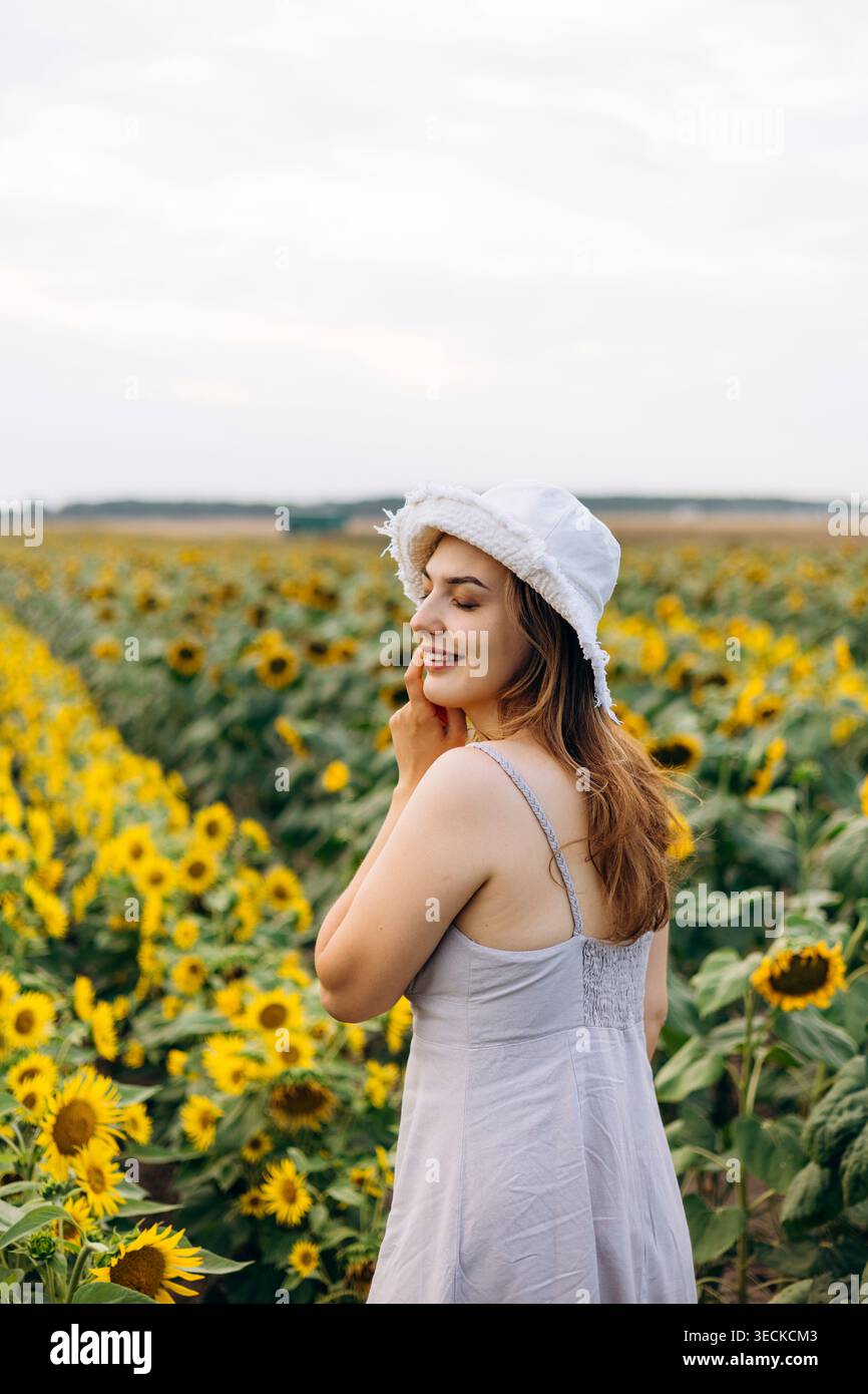 Die lächelnde junge Frau mit weißem Hut und Sommerkleid steht zur goldenen Stunde auf einem blühenden Sonnenblumenfeld und genießt die Natur und die ruhige ländliche Atmosphäre Stockfoto