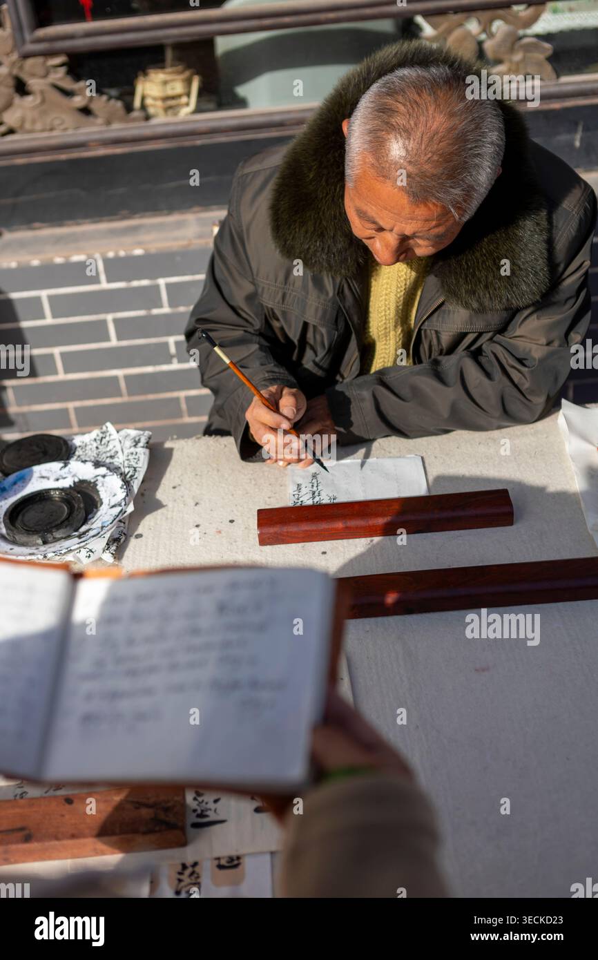 Ein älterer Mann übt traditionelle chinesische Kalligraphie mit Pinsel und Tinte an einem Tisch im Freien und schreibt Schriftzeichen im Winterlicht auf Papier, Konservi Stockfoto