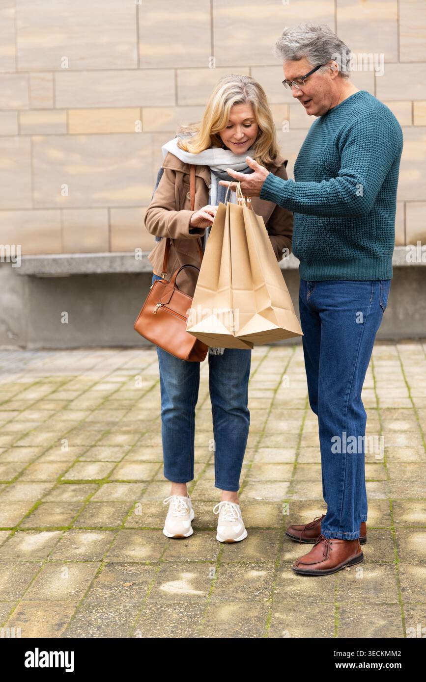 Seniorenpaar, das auf dem gepflasterten platz steht, hält braune Papiertaschen mit brauner Ledertasche Stockfoto
