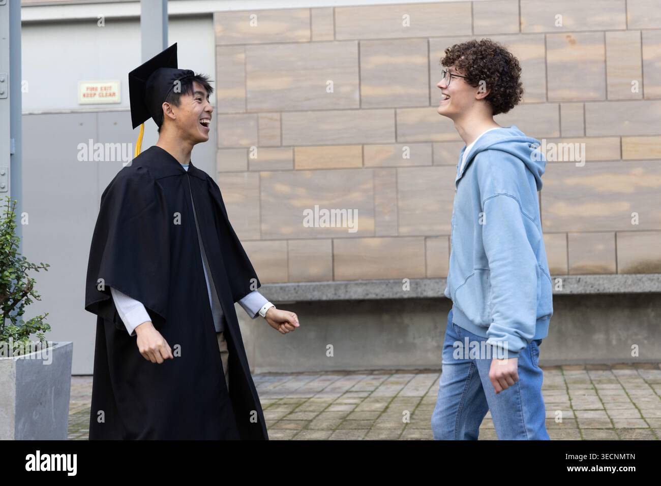 Verschiedene männliche Freunde, einer im Hoodie, feiert den Abschluss im Kleid, Hut mit gelber Quaste im Innenhof Stockfoto