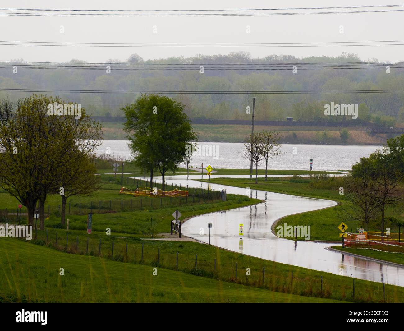 Kurvige Straße durch grünen Park an einem regnerischen Frühlingstag mit nassem Bürgersteig, der das Licht in der Nähe eines ruhigen Sees und Bäumen reflektiert Stockfoto