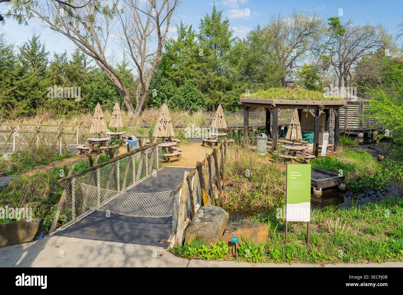 Eine hölzerne Fußgängerbrücke überquert einen kleinen Bach in Richtung eines schattigen Essbereichs im Freien mit Sonnenschirmen und Picknicktischen unter Bäumen. Stockfoto