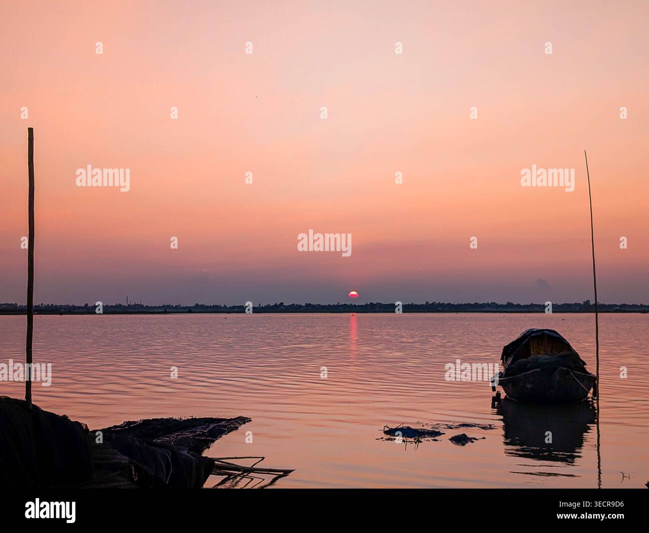 Friedlicher Sonnenuntergang am Fluss mit einem kleinen hölzernen Boot vor einem warmen orangen Himmel, das sich auf ruhigem Wasser im ländlichen Bangladesch reflektiert. - Smartphone-aufgenommenes Stockfoto