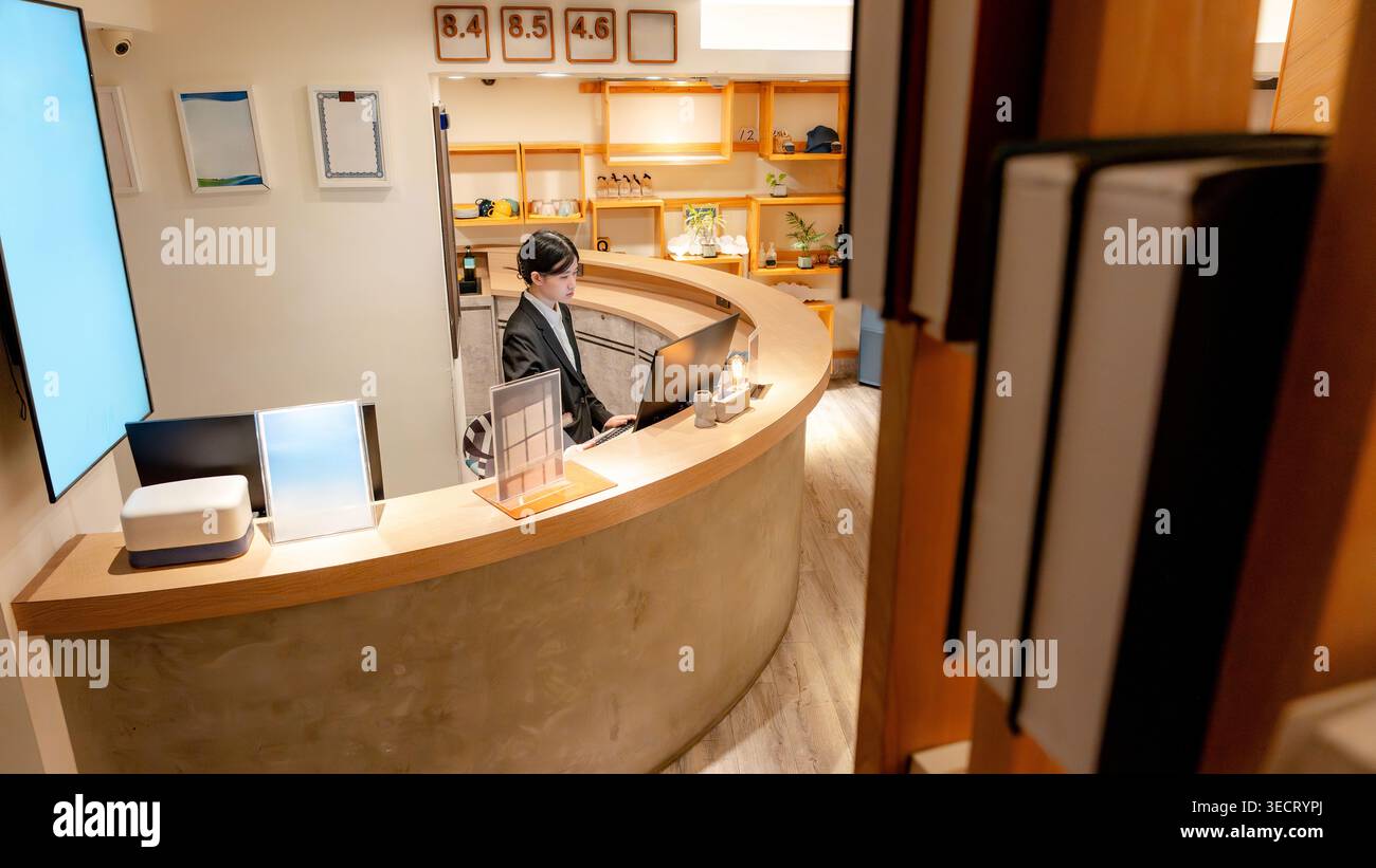 Der Rezeptionist des taiwanesischen Hotels arbeitet an einer modernen, geschwungenen Rezeption und nutzt einen Computer in einer warmen, minimalistischen Lobby, der den Gästen beim Check-in hilft Stockfoto