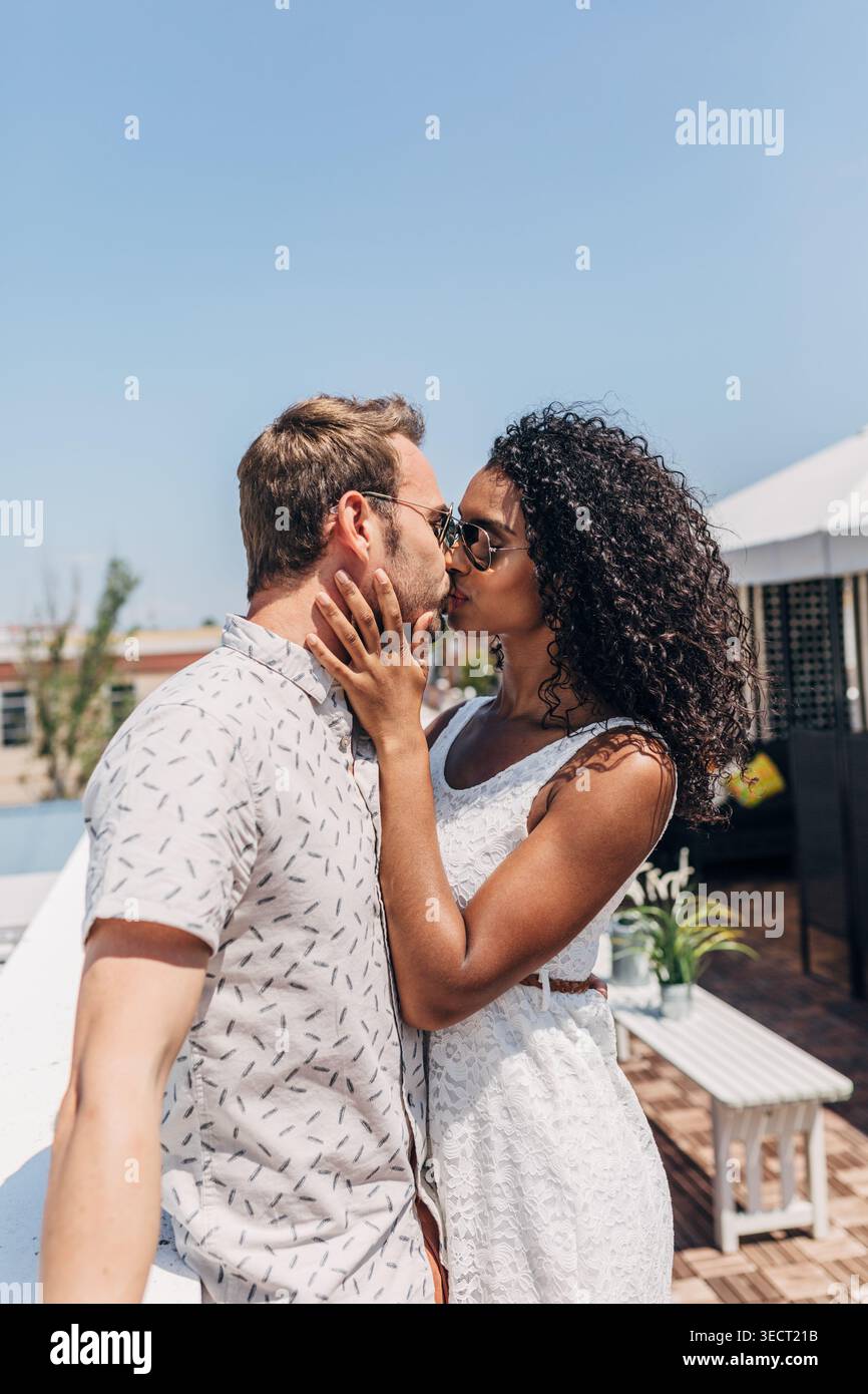 Romantisches Paar küsst sich an einem sonnigen Sommertag im Freien, trägt Sonnenbrille in lässigen Outfits, feiert Liebe und Intimität auf einer hellen Terrasse mit c Stockfoto