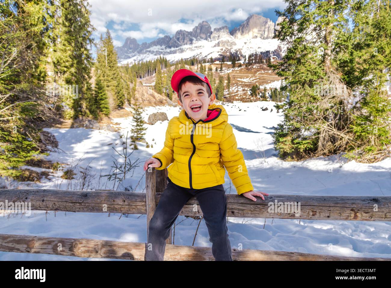 Lächelndes Kind in gelber Jacke, das im Winter auf einem Holzzaun in der Nähe des Karersee sitzt, umgeben von Schnee, Kiefernwald und Dolomitengipfeln im Alto Adi Stockfoto