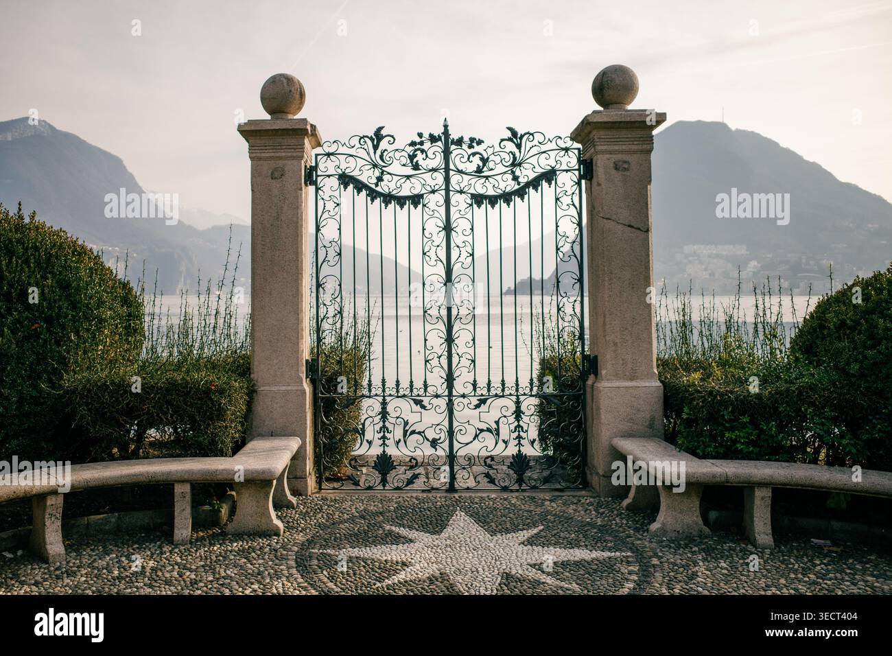 Ein kunstvoll verziertes schmiedeeisernes Tor öffnet sich zu einem atemberaubenden Blick auf einen ruhigen europäischen See, der von majestätischen Bergen umgeben ist und das Wesen von Reise und wander verkörpert Stockfoto