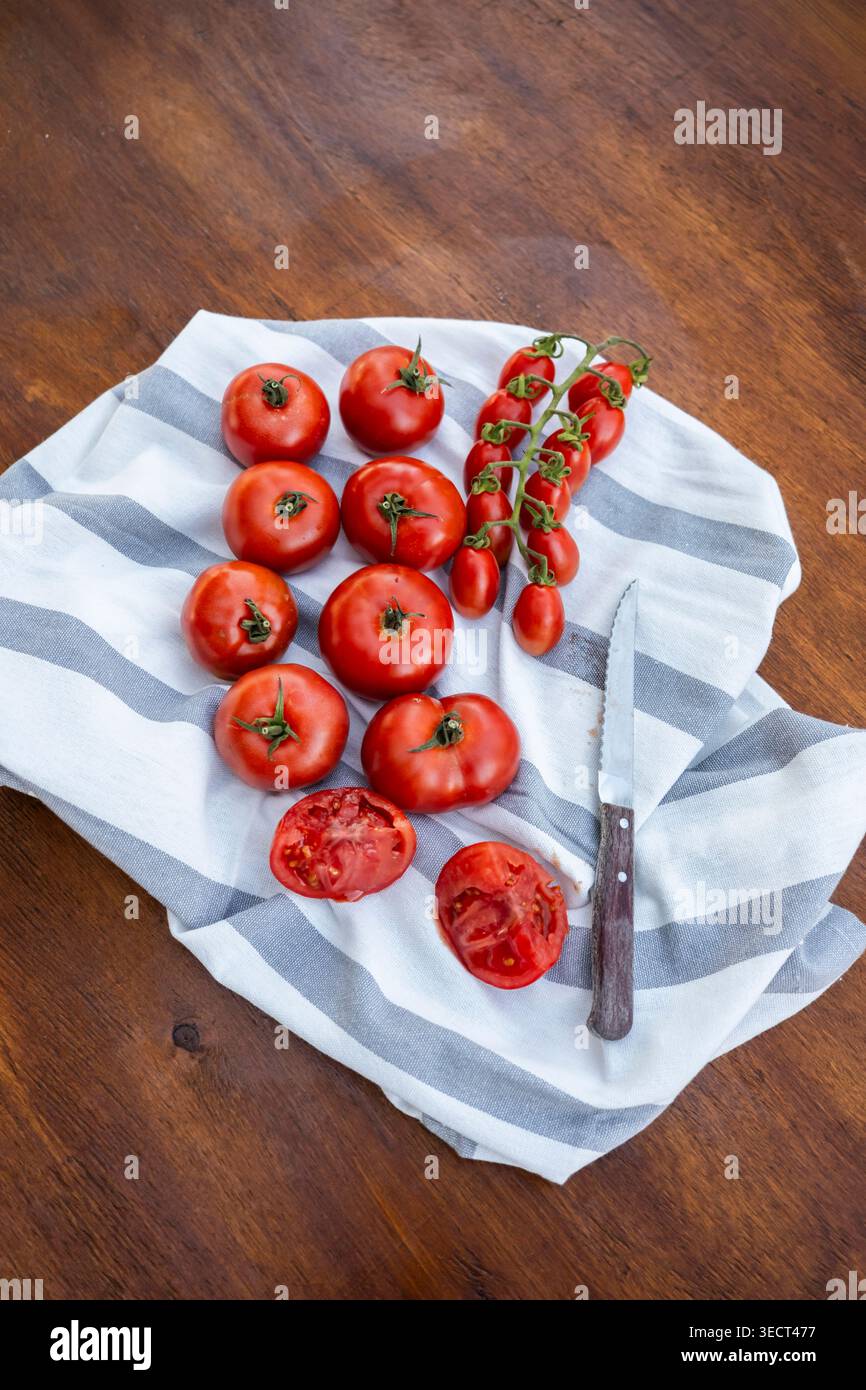 Blick von oben auf reife rote Tomaten, einschließlich Kirschtomaten auf der Weinrebe, auf einem gestreiften Küchentuch auf einem rustikalen Holztisch mit einem Messer und sl angeordnet Stockfoto