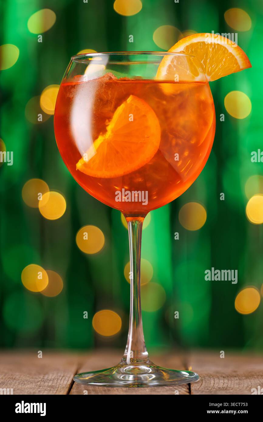 Aperol spritzt Cocktail mit Orangenscheibe und Eis auf rustikalem Holztisch, festliche grüne und goldene Bokeh-Lichter Stockfoto