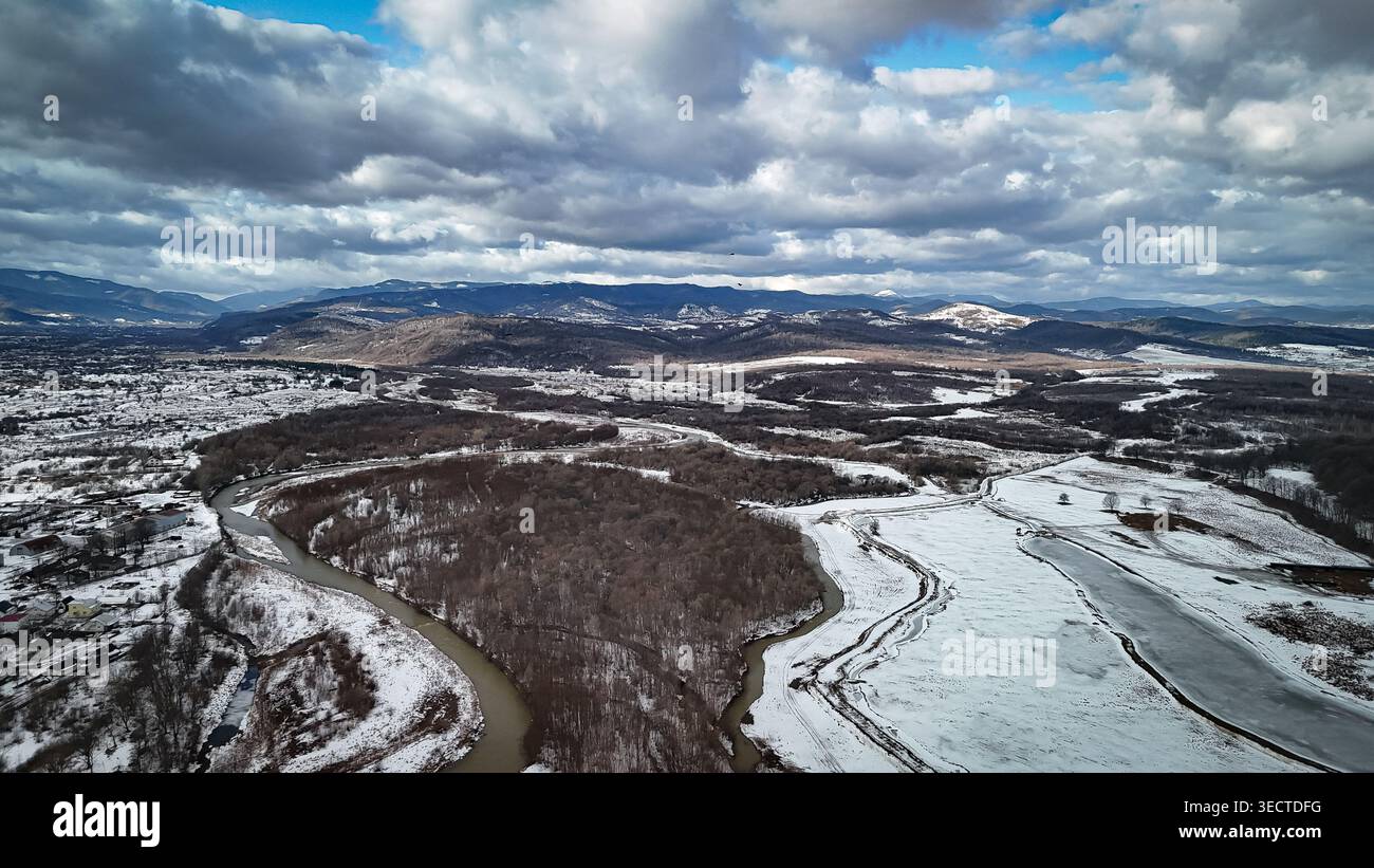 Die Luftperspektive zeigt ein winterliches Tal aus schlängelnden Flüssen, Wäldern und schneebedeckten Feldern, die von dunklen Bergrücken eingerahmt werden. Schwere Wolken ziehen über sich Stockfoto