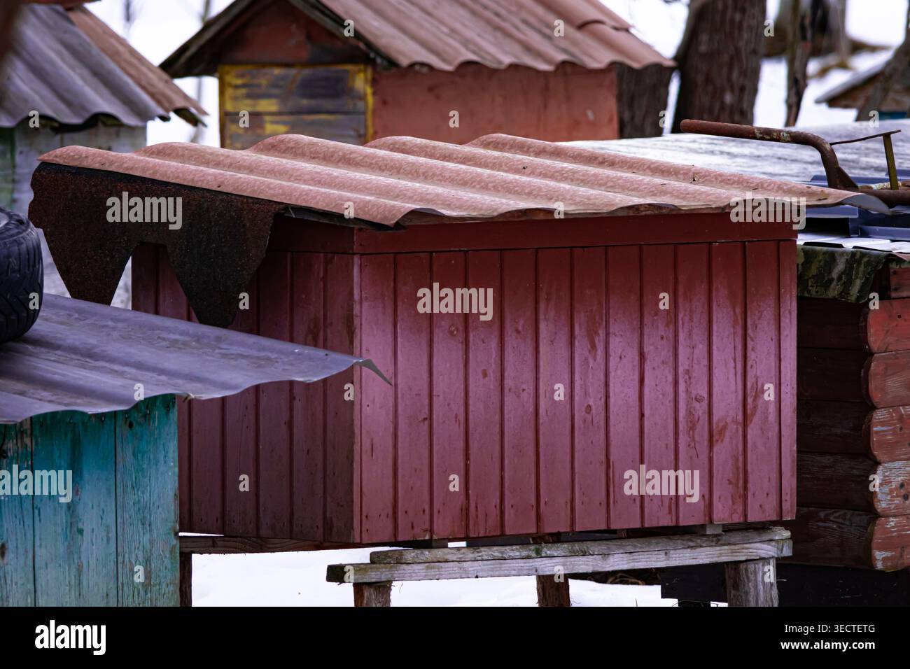 Nahporträt eines rotbraunen Bienenstöckchens mit Zinndach. Schneebänke umgeben die Struktur, während gedämpftes Licht Korn und Knoten in den Dielen freigibt Stockfoto