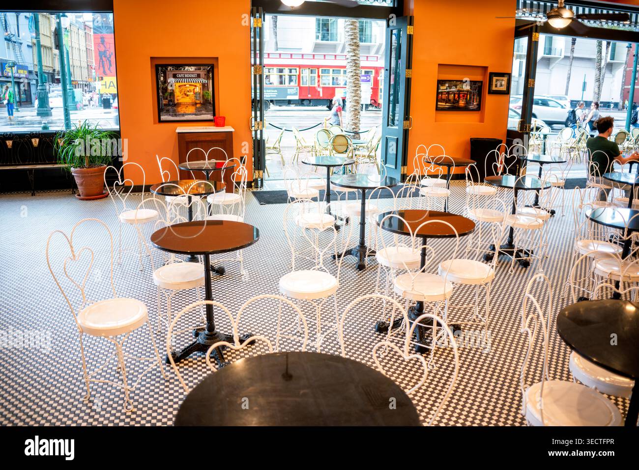 Cafe Beignet Canal Street Karierter Boden Drahtstühle New Orleans Vereinigte Staaten | NEW ORLEANS, Louisiana — Weiße Drahtstühle und dunkle Sockeltische ruhen auf einem schwarz-weißen karierten Boden im Cafe Beignet, wo offene Türen einen roten Straßenbahnwagen zeigen, der die Canal Street entlangfährt. Das Innendesign, das mit herzförmigen Bistrotischen und geometrischem Fliesenwerk punktet, orientiert sich an der traditionellen Ästhetik französischer Cafés aus dem späten 19. Jahrhundert und spiegelt das tiefe frankophone Erbe der Stadt wider. Jenseits der Schwelle verkehrt die Canal Street Straßenbahnlinie mit markanten roten Wagen, die modern und klimatisiert sind Stockfoto