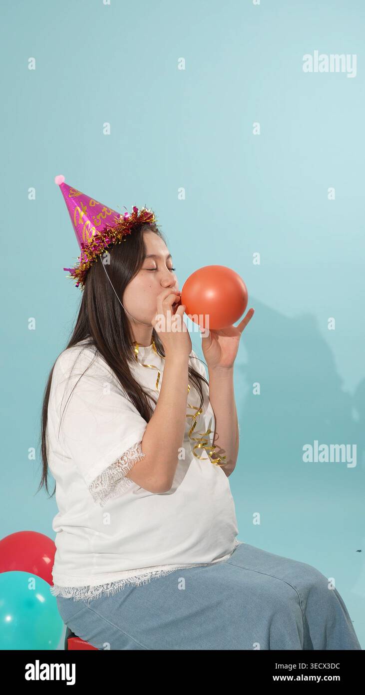 Schwangere Frau mit Partyhut, die einen orangen Ballon in die Luft jagt Stockfoto