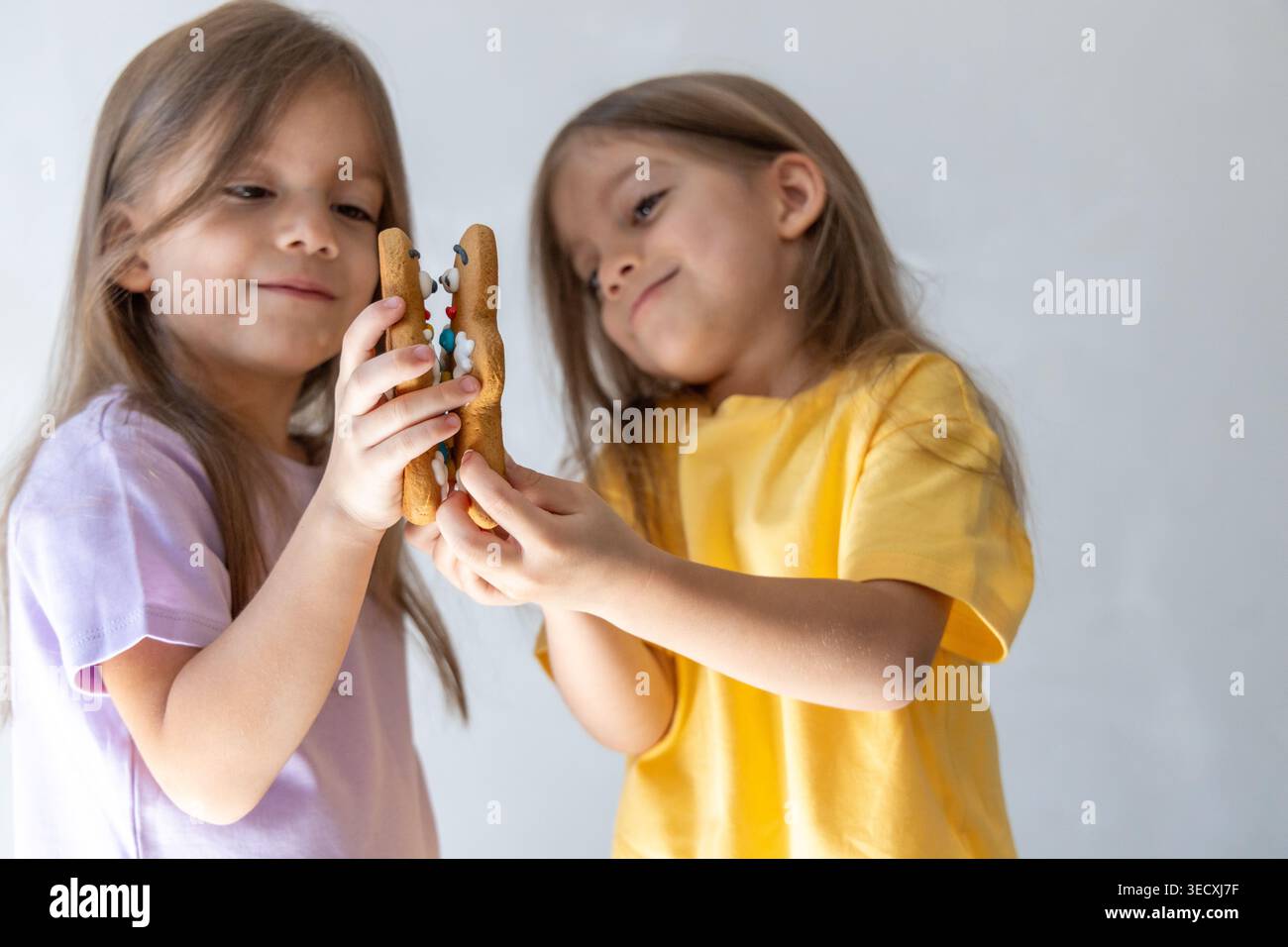 Zwei Schwestern Lächeln, Während Sie Festliche Kekse Austauschen. Zwei Mädchen In Gelb Und Lavendel, Die Süße Lebkuchenspielzeug Teilen Stockfoto