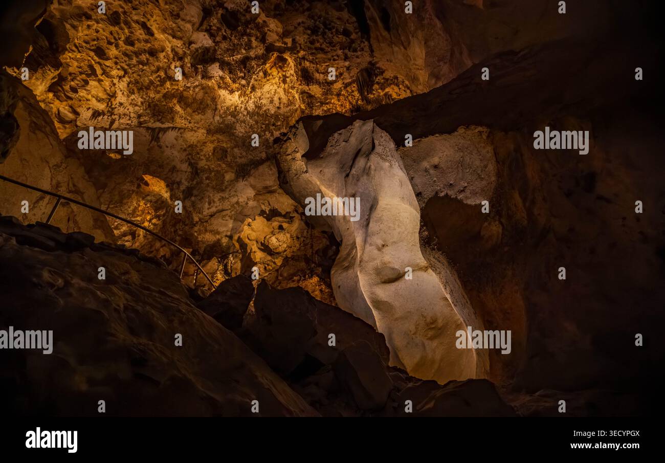 Helles Sonnenlicht hebt strukturierte braune Felswände hervor und einen hoch aufragenden weißen Stalagmiten, der sich in der Nähe eines dunklen Treppengeländes in einer natürlichen Höhle erhebt. Stockfoto