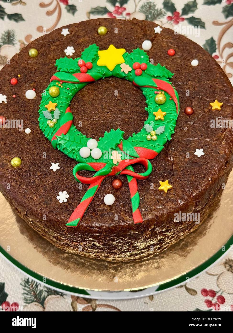 Ein traditioneller dunkler Weihnachts-Fruchtkuchen, dekoriert mit einem essbaren grünen Fondant-Weihnachtskranz, Zuckersternen und festlichen Streuseln auf einer goldenen Kuchenplatte. - Smartphone-aufgenommenes Stockfoto