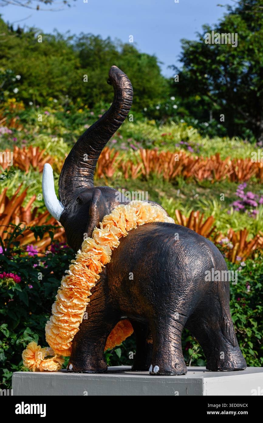 Schnitzerei eines Elefanten, ein Symbol für Stärke, Geduld, Loyalität und Weisheit, mit einer orangen Blumengirlande bedeckt, neben einem kleinen buddhistischen Tempel auf Thai Stockfoto