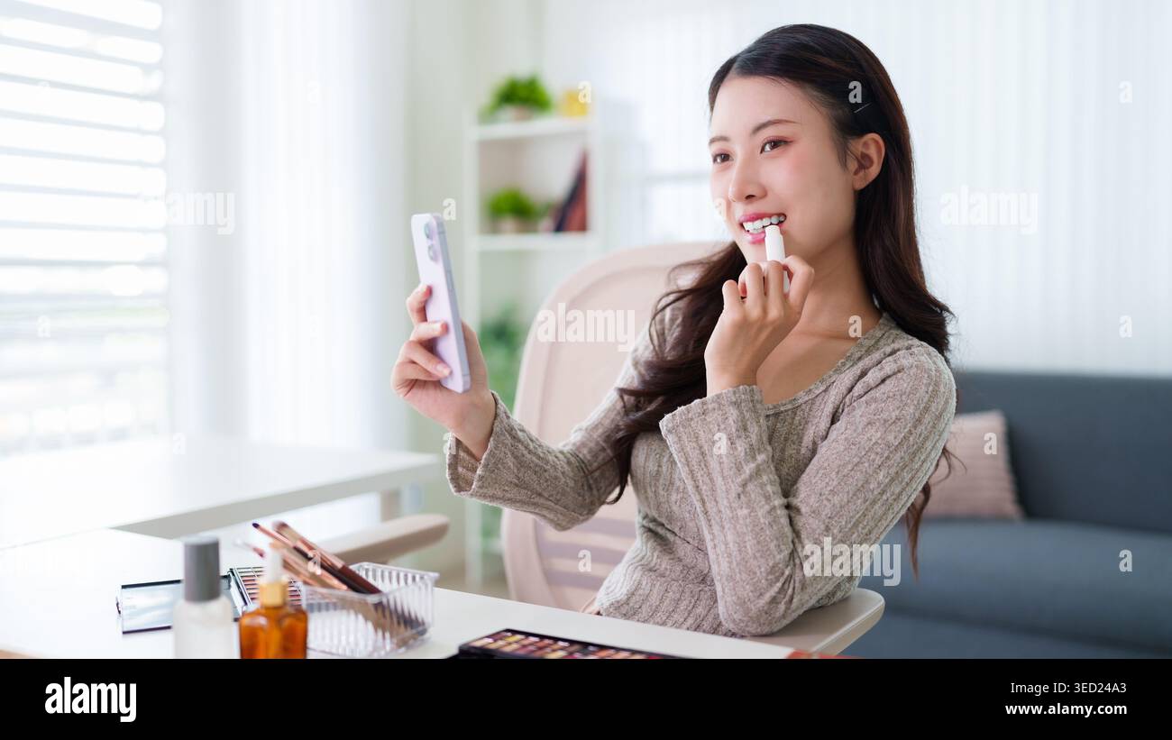 Junge asiatische Frau, die Lippenstift anwendet und auf den Smartphone-Spiegel schminkt, die Schönheitsroutine zu Hause im Wohnzimmer macht Stockfoto
