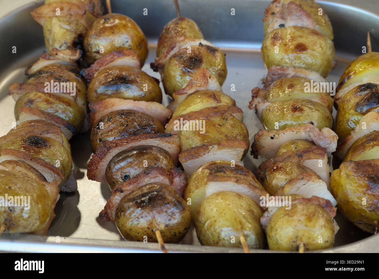 Saftige Kartoffeln, in knusprigem Speck gewickelt, brutzeln während eines sonnigen Kochens auf Spießen. Stockfoto