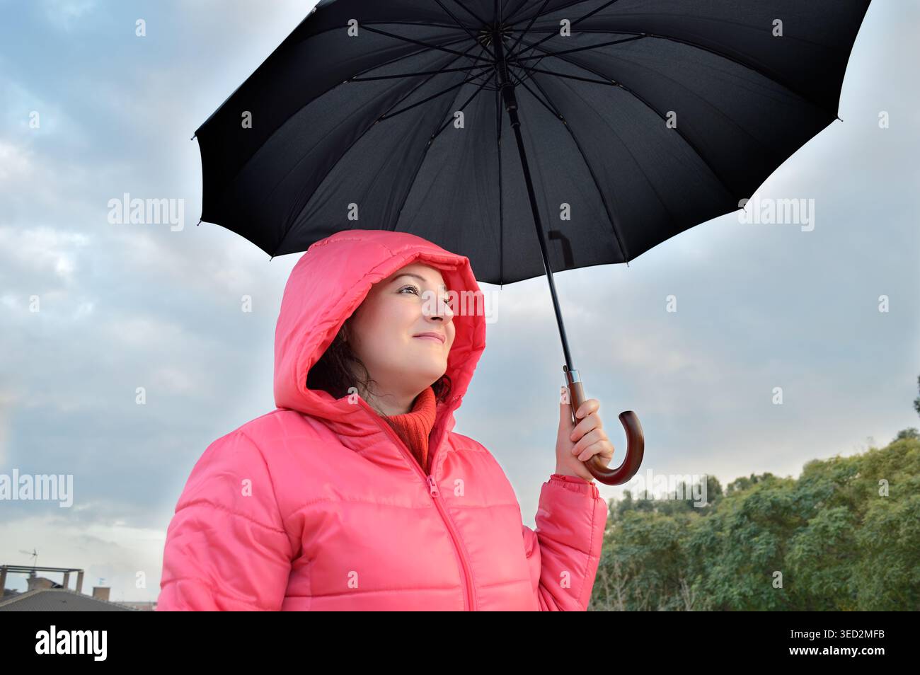 Schöne junge Frau, die einen rosafarbenen Anorak trägt, hält im Herbst einen schwarzen Regenschirm, überrascht und aufgeregt unter dem angehaltenen Regen Stockfoto