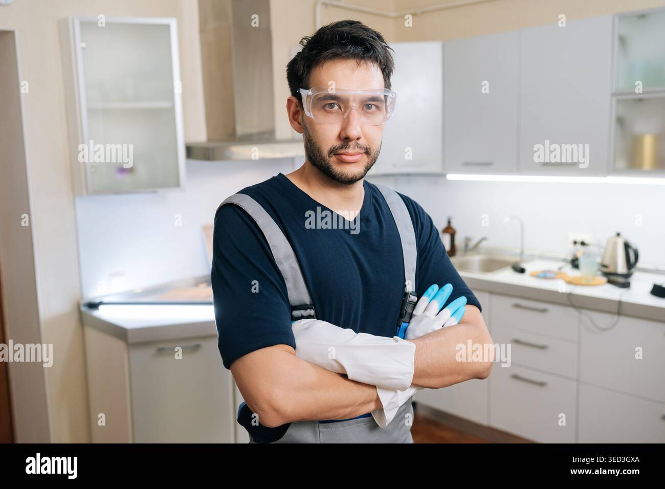 Professioneller männlicher Arbeiter mit Schutzbrille und Arbeitshandschuhen, überkreuzte Arme, während er selbstbewusst in der modernen Küche steht, bereit für die Reinigung Stockfoto