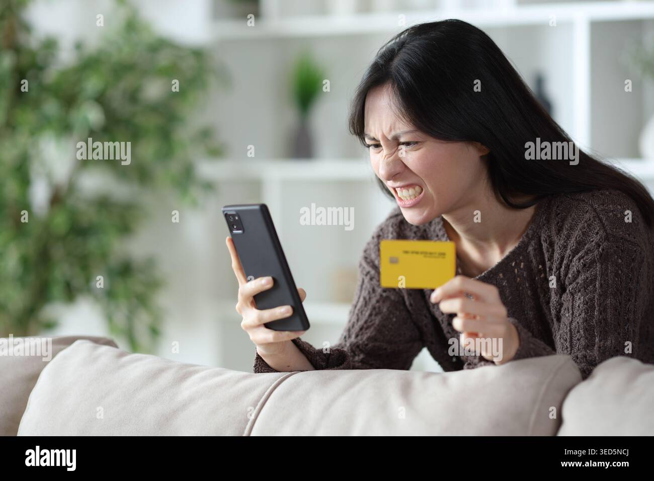 Wütende asiatische Frau bezahlt online mit Kreditkarte und Telefon zu Hause Stockfoto
