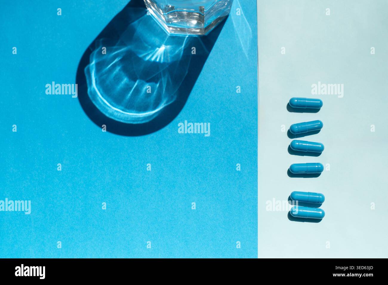 Blaue pharmazeutische Kapseln in der Nähe eines Wasserglases in einem Atelier verstreut. Konzept der täglichen Vitaminaufnahme, des Behandlungsprozesses und der hohen Qualität Stockfoto