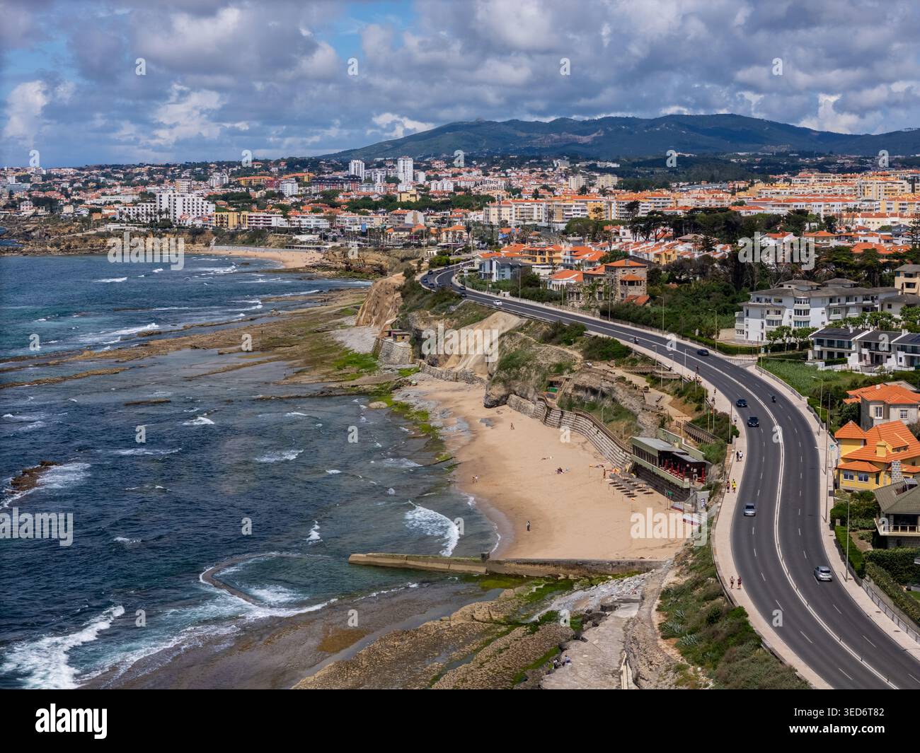 Aus der Vogelperspektive auf die Avenida Grenzstraße entlang der felsigen Küste und dem Sandstrand mit Wohngebäuden und Hügeln unter einer bewölkten Landschaft Stockfoto