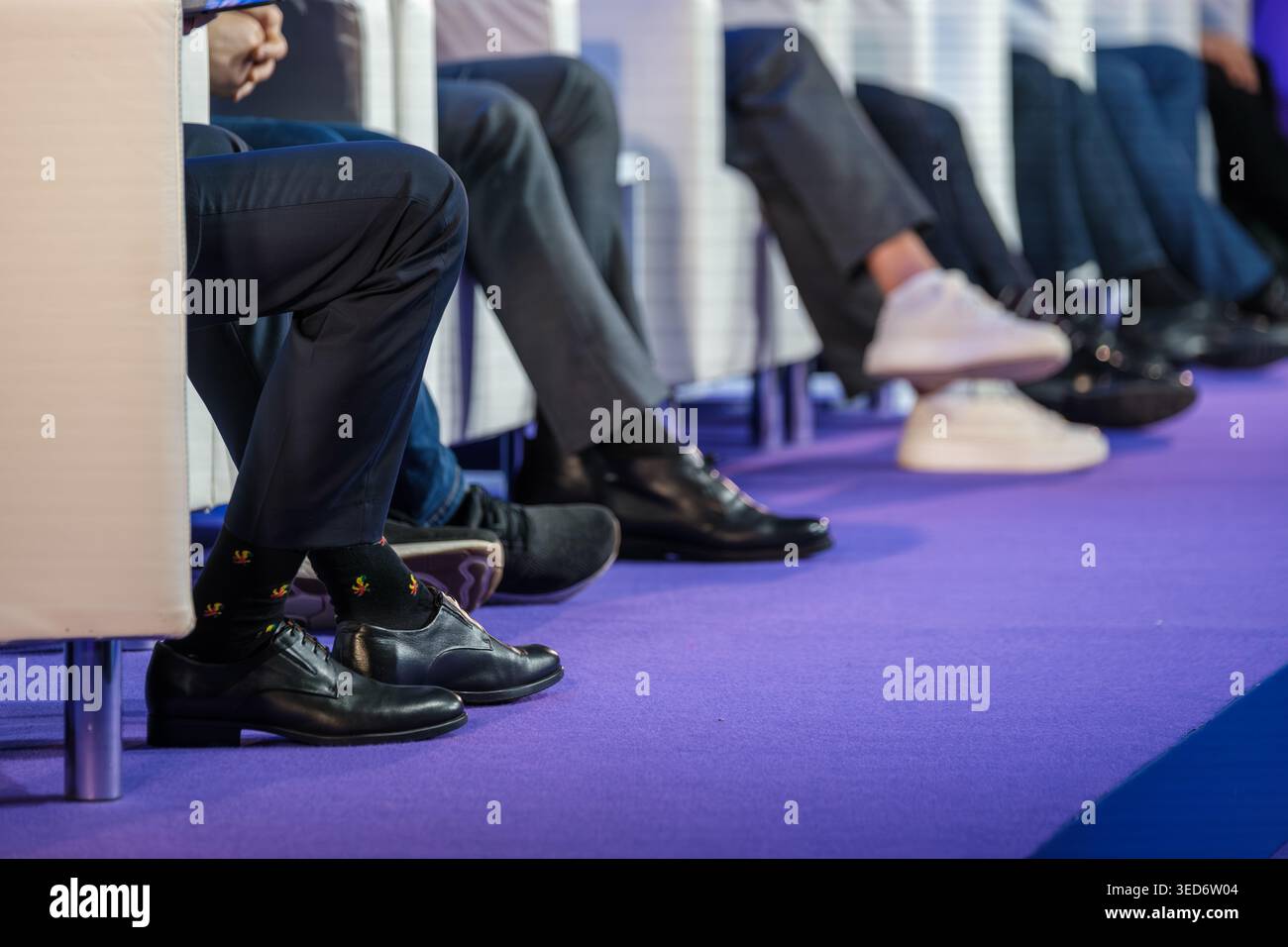 Auf der Bühne sitzen die Diskussionsteilnehmer, die sich auf Beine und Schuhe konzentrieren, den Kontrast zwischen Kleidern und Turnschuhen, die Atmosphäre eines formellen Treffens. Stockfoto