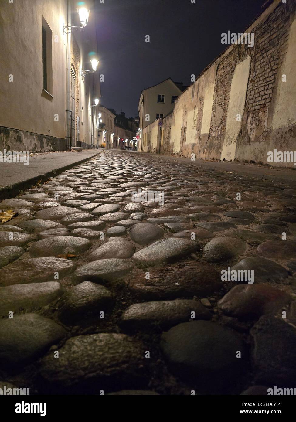 Eine enge Kopfsteinpflasterstraße, die nachts von Straßenlaternen beleuchtet wird, in der Altstadt von Vilnius, Litauen - Smartphone-aufgenommenes Stockfoto