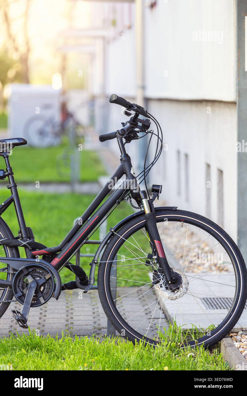 Schwarzes Fahrrad geparkt Wohnbaugebiet grüner Rasen gepflasterter Fußweg ruhiges Stadtviertel Deutschland. Moderner Fahrradkorb für Stadtfahrräder steht für Sonnenlicht Stockfoto