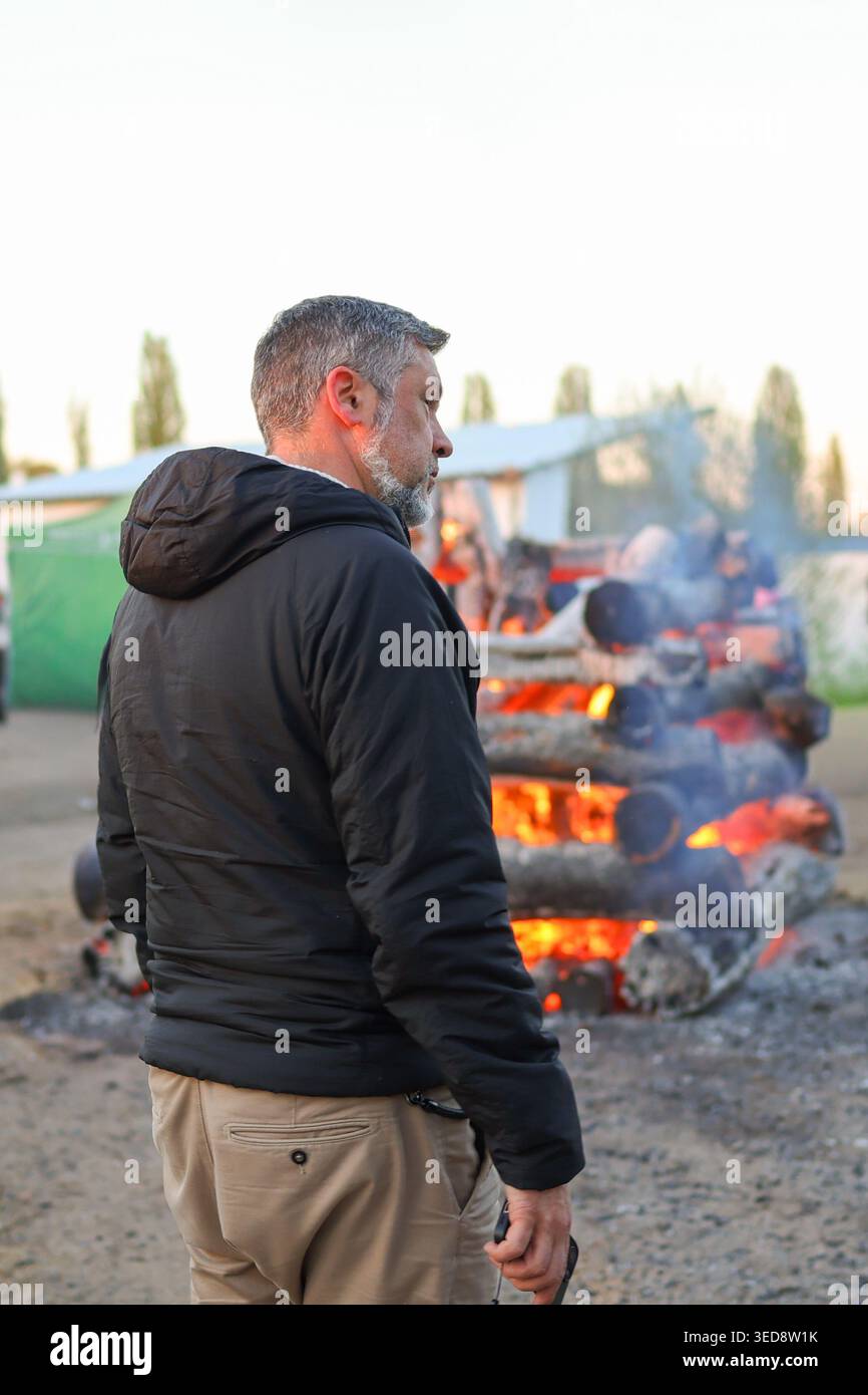 Ein glücklicher Mann mittleren Alters posiert bei einer Veranstaltung im Freien am brennenden Holzfeuer und fängt die gemütliche Atmosphäre und den saisonalen Lifestyle-Moment ein. Stockfoto