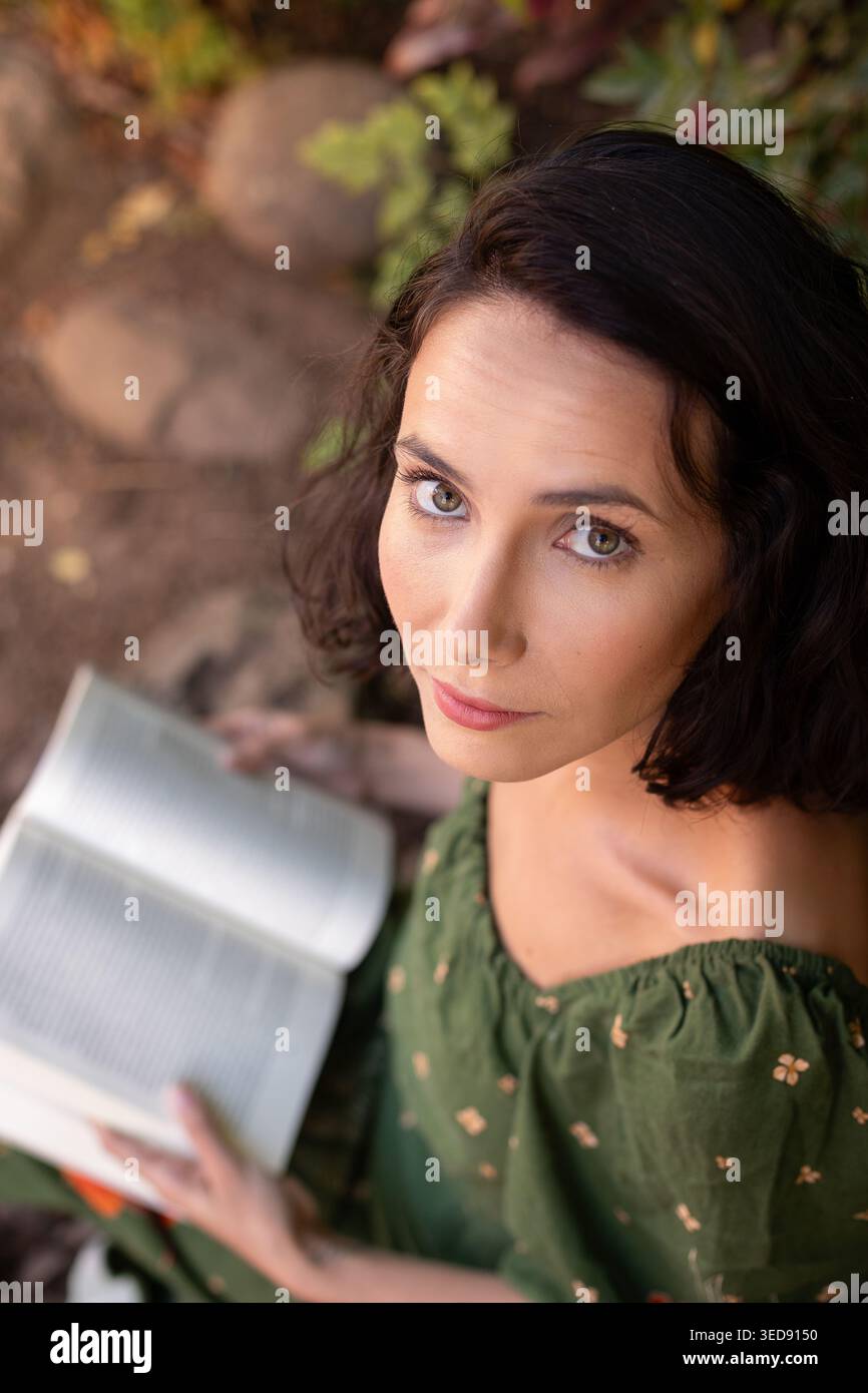 Eine Frau mit kurzen Haaren sitzt auf dem Boden mit einem Buch in der Hand. Sie scheint konzentriert und engagiert zu sein, während sie von Grün umgeben ist. Das Sonnenlicht leuchtet Stockfoto