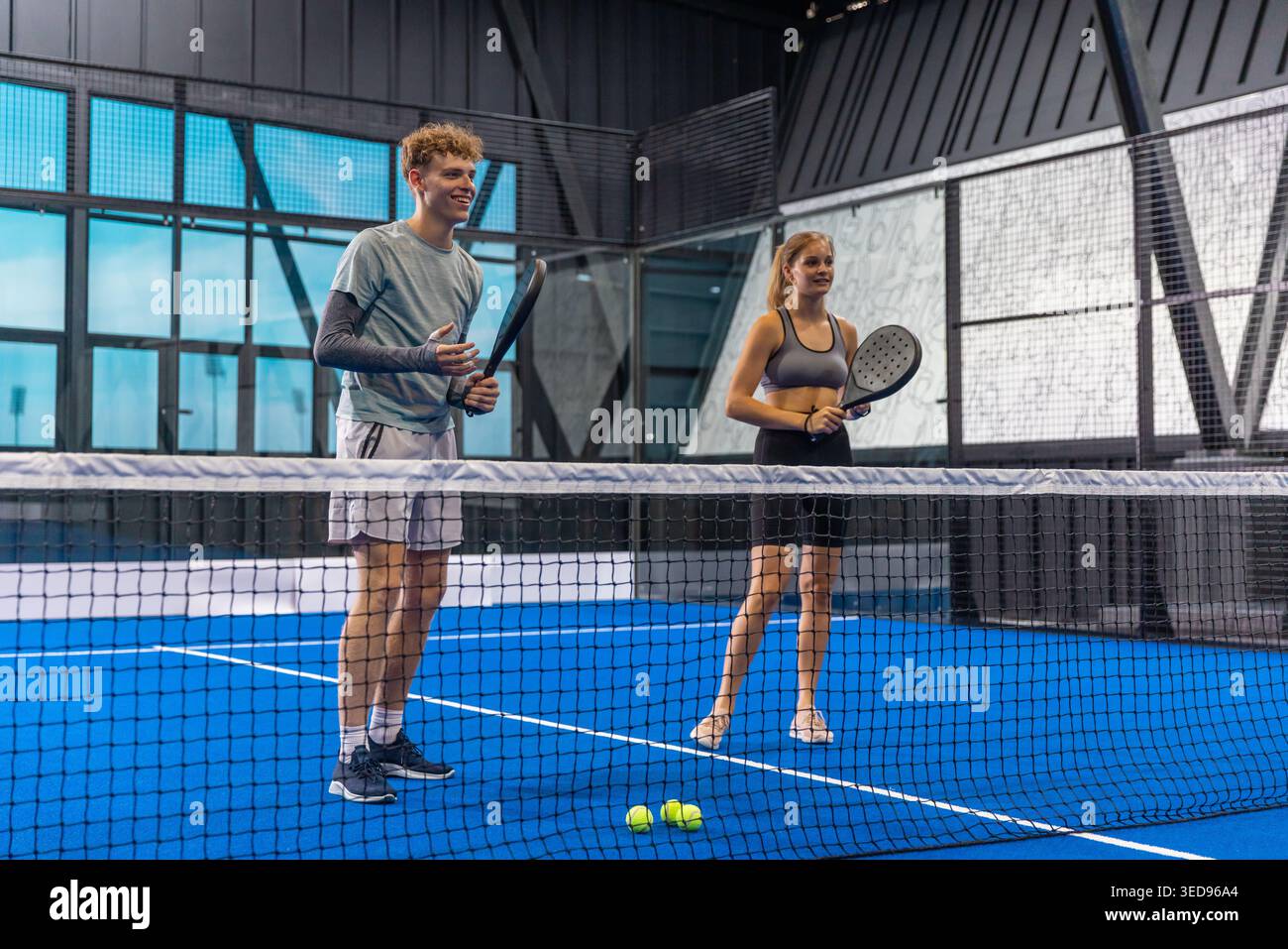 Freunde stehen hinter dem Netz auf dem blauen Padelplatz mit getönten Fenstern, die Schläger und Bälle halten Stockfoto