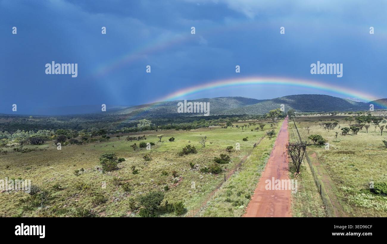 Aus der Vogelperspektive einer roten Feldstraße, die sich durch die grüne Savannenlandschaft unter einem doppelten Regenbogen und dunklem stürmischen Himmel erstreckt. Waterberg District Municipali Stockfoto