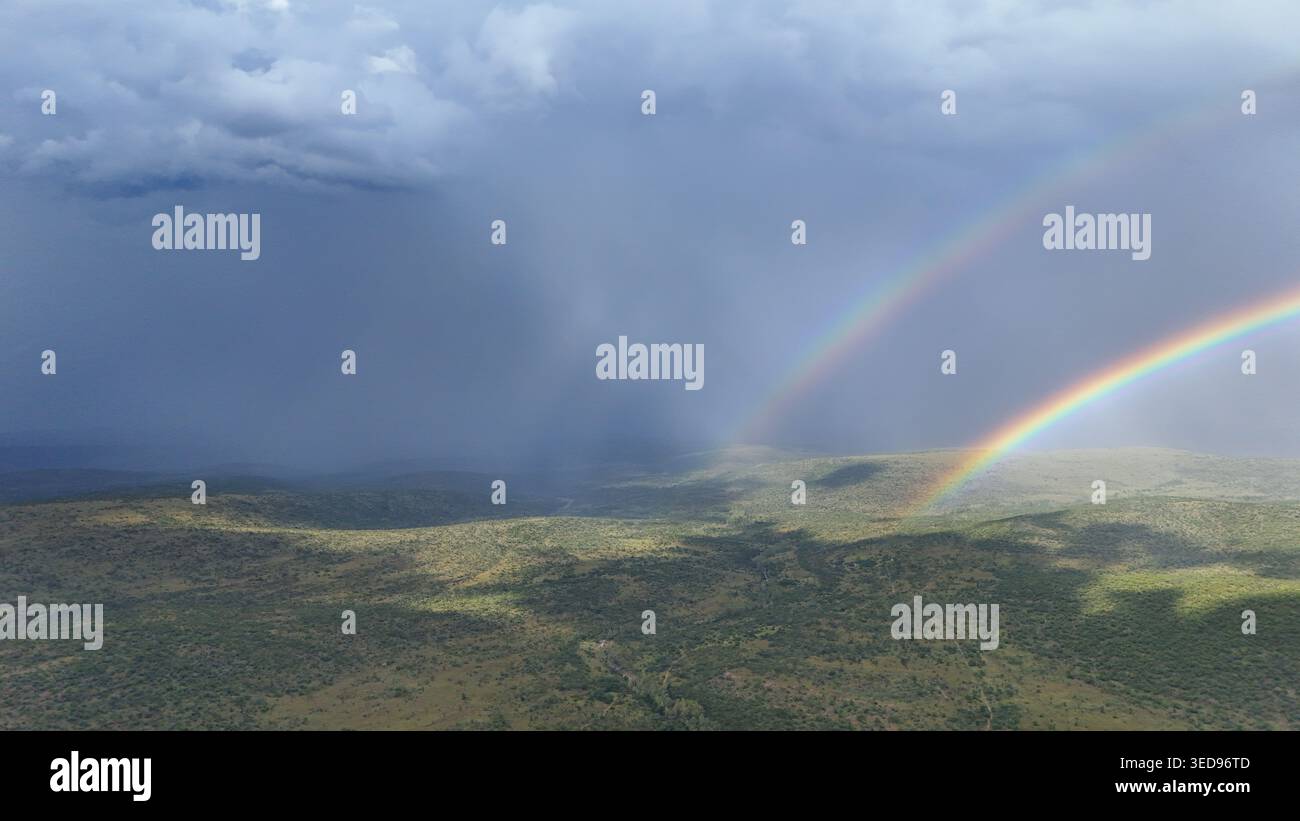 Luftaufnahme eines Doppelregenbogens, der sich über eine weite grüne Landschaft unter dunklen Sturmwolken in Waterberg District Municipality, Limpopo, South AF erstreckt Stockfoto