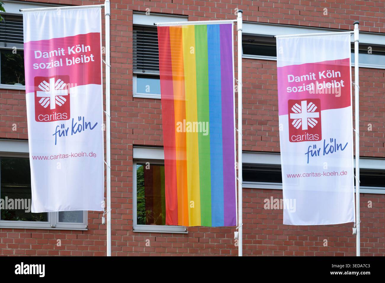 Köln, Deutschland 02. Juni 2025: Drei Fahnen hängen an Pfählen vor einem roten Backsteingebäude: Zwei Fahnen der Caritas „für köln“ und ein vertikaler Regenbogenstolz Stockfoto
