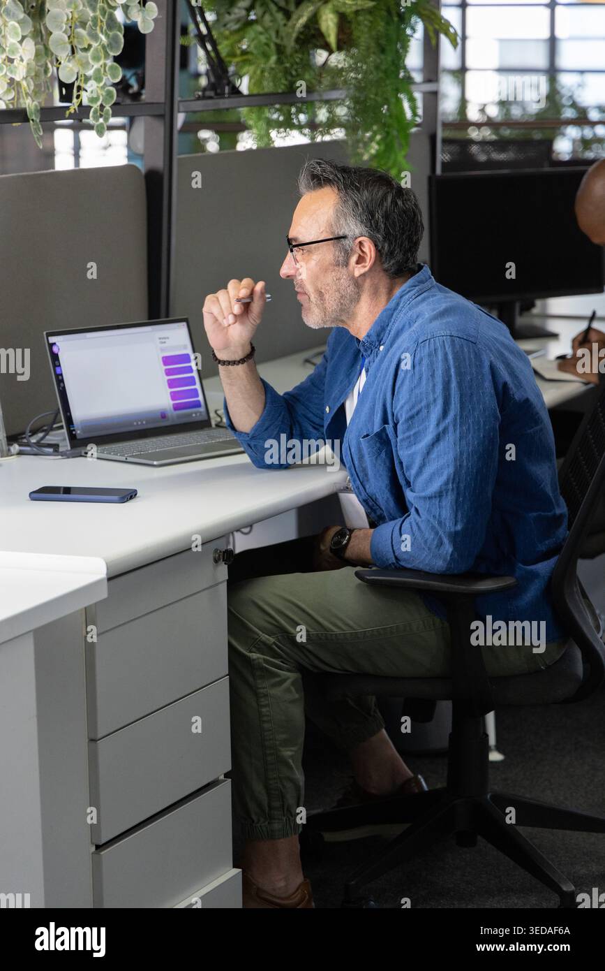 Verschiedene Kollegen, die im Open Office arbeiten; Mann in blauem Hemd, der Laptop-violetten Chat und Telefon überprüft Stockfoto