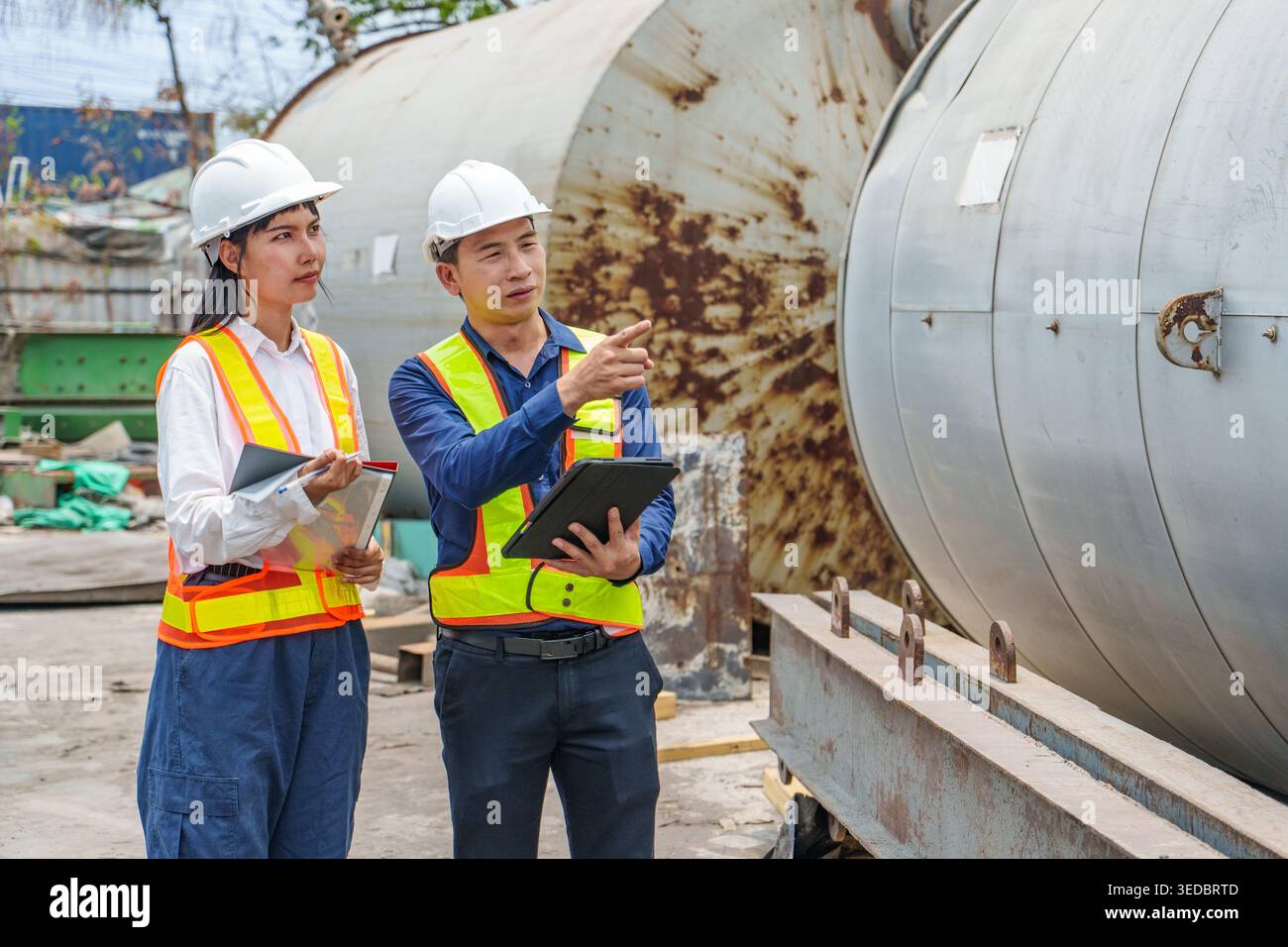 Professionelles Industrieteam in Hochsichtwesten, das Daten auf einem Tablet überprüft und während einer Inspektion auf große Metalltanks zeigt Stockfoto