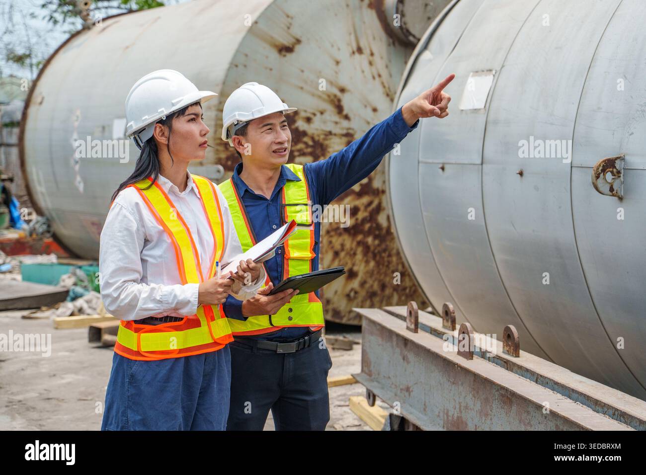 Professionelles Industrieteam in Hochsichtwesten, das Daten auf einem Tablet überprüft und während einer Inspektion auf große Metalltanks zeigt Stockfoto