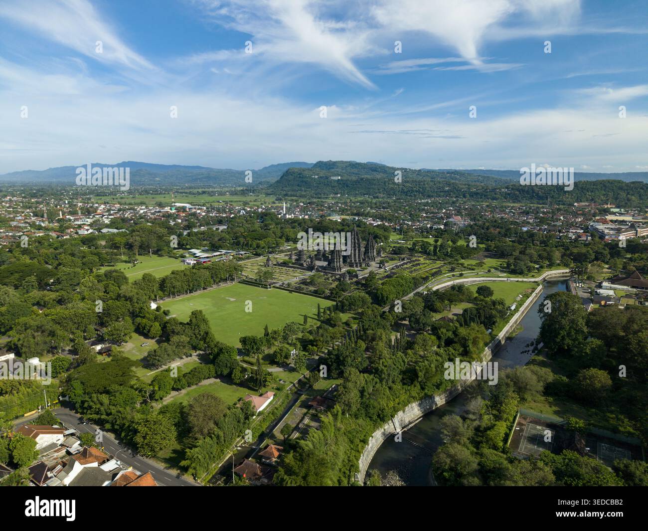 Blick aus der Vogelperspektive auf den Prambanan Temple Complex, umgeben von üppigen grünen Feldern, einem sich windenden Fluss und der städtischen Landschaft unter einem blauen Himmel mit schimmernden Wolken in PR Stockfoto
