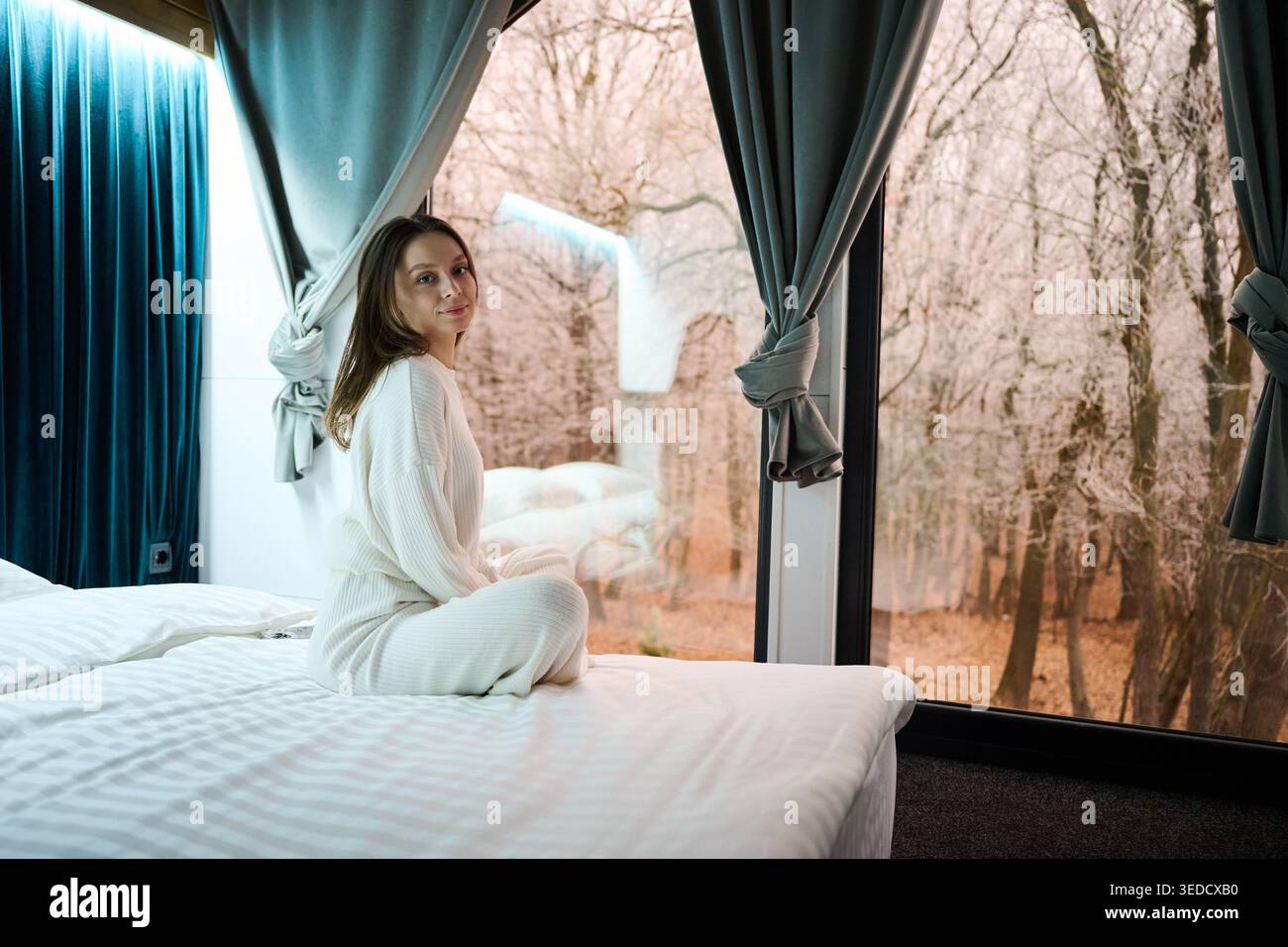 Junge Frau, die sich auf dem Bett in der Nähe des Panoramafensters entspannt, mit frostigen Bäumen draußen, friedliche Wohnumgebung, Winterstimmung, Komfort, langsames Leben, und ein ruhiger Indoor-Lifestyle Stockfoto