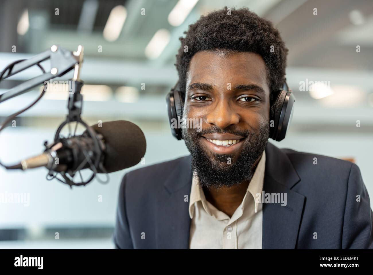 Happy man trägt Kopfhörer und einen dunklen Anzug, der digitale Audioinhalte in einem professionellen Studio erstellt und seine Stimme mit einer Audioanlage für eine Online-Show überträgt Stockfoto