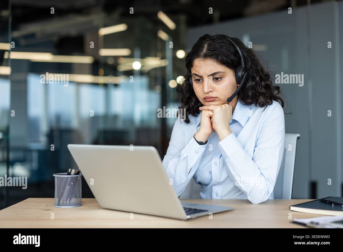 Gestresste junge Frau im Headset an einem modernen Bürotisch, starrte mit Erschöpfung und Frustration auf den Laptop, erlebte Burnout und Enttäuschung während der Fernarbeit Stockfoto