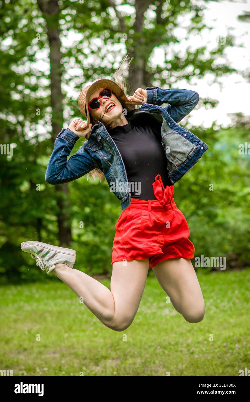 Mädchen mit Strohhut, Jeansjacke, roten Shorts und weißen Sneakers mit roter Herzsonnenbrille und leuchtend rotem Lippenstiftsprung. Stockfoto