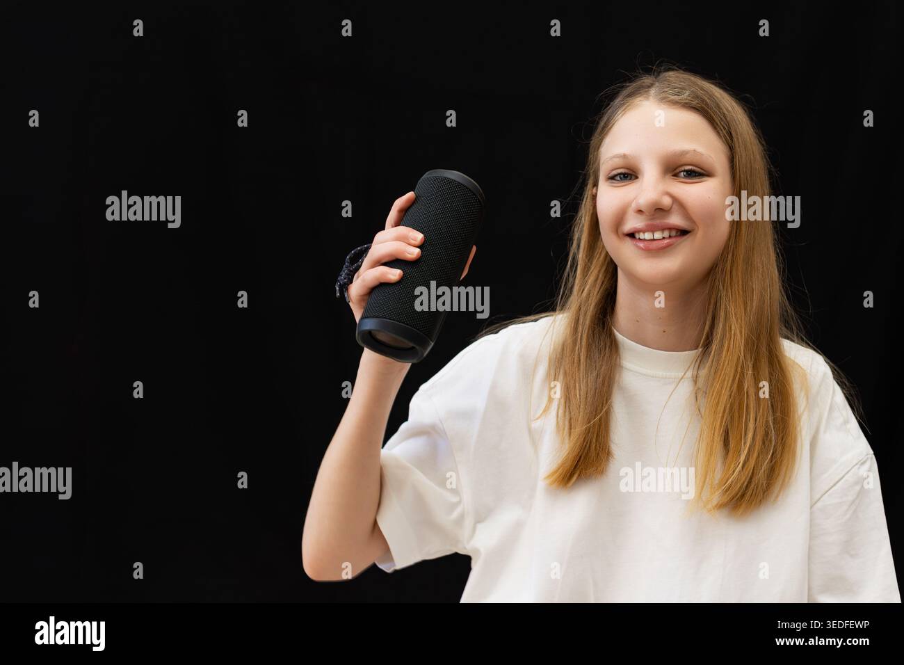 Lächelndes Teenager-Mädchen, Das Einen Tragbaren Lautsprecher Hält. Glückliches blondes Mädchen, das einen schwarzen kabellosen Lautsprecher hält, der Musik und Audiotechnologie repräsentiert Stockfoto
