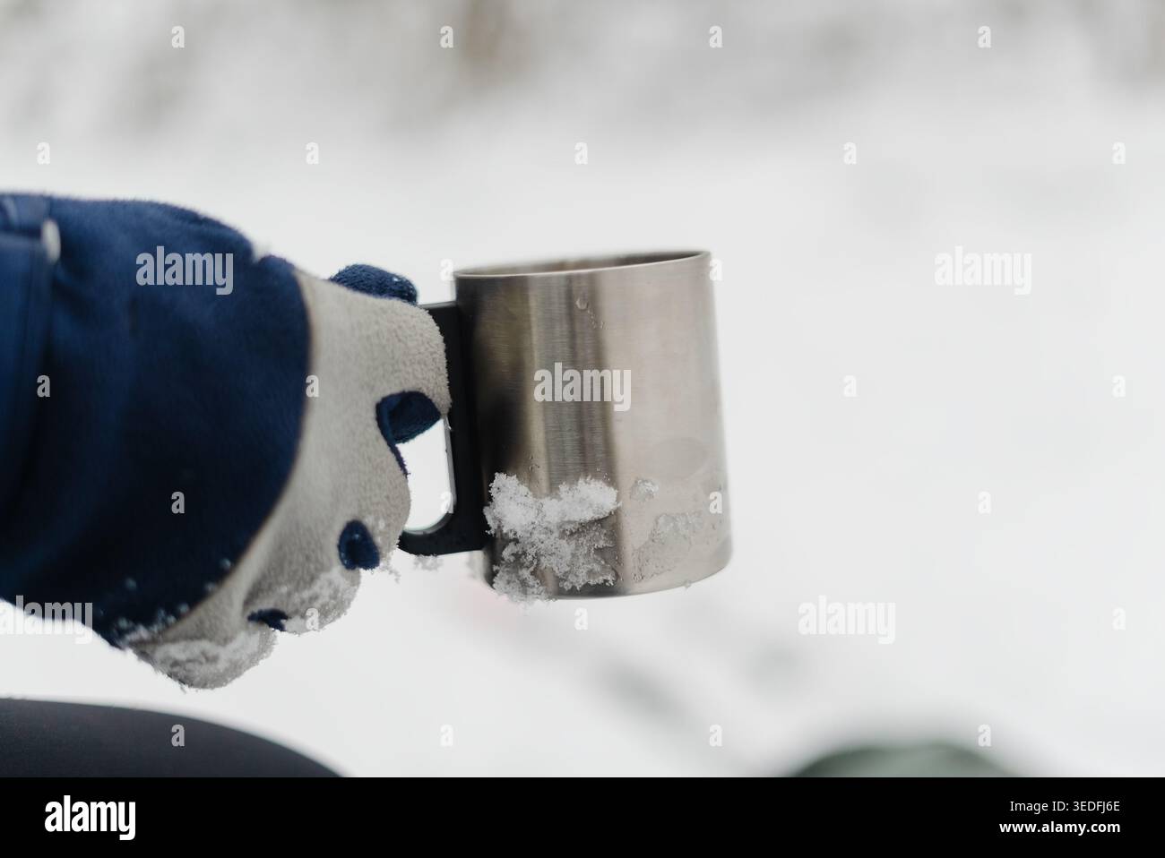 Handschuhe greifen eine Metallschale im Winter draußen. Blau-weißer Winterhandschuh mit Thermobecher für ein Heißgetränk bei eiskaltem Wetter. Praktische Outdoor-Ausrüstung für kaltes Wetter an einem verschneiten Tag Stockfoto